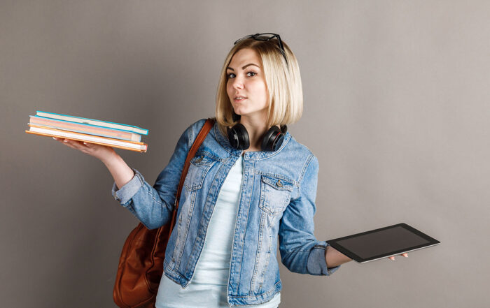 Book vs. e-book. Girl student holding a traditional textbook and reading e-books isolated on gray background. Choose between paper books and e-learning gadgets