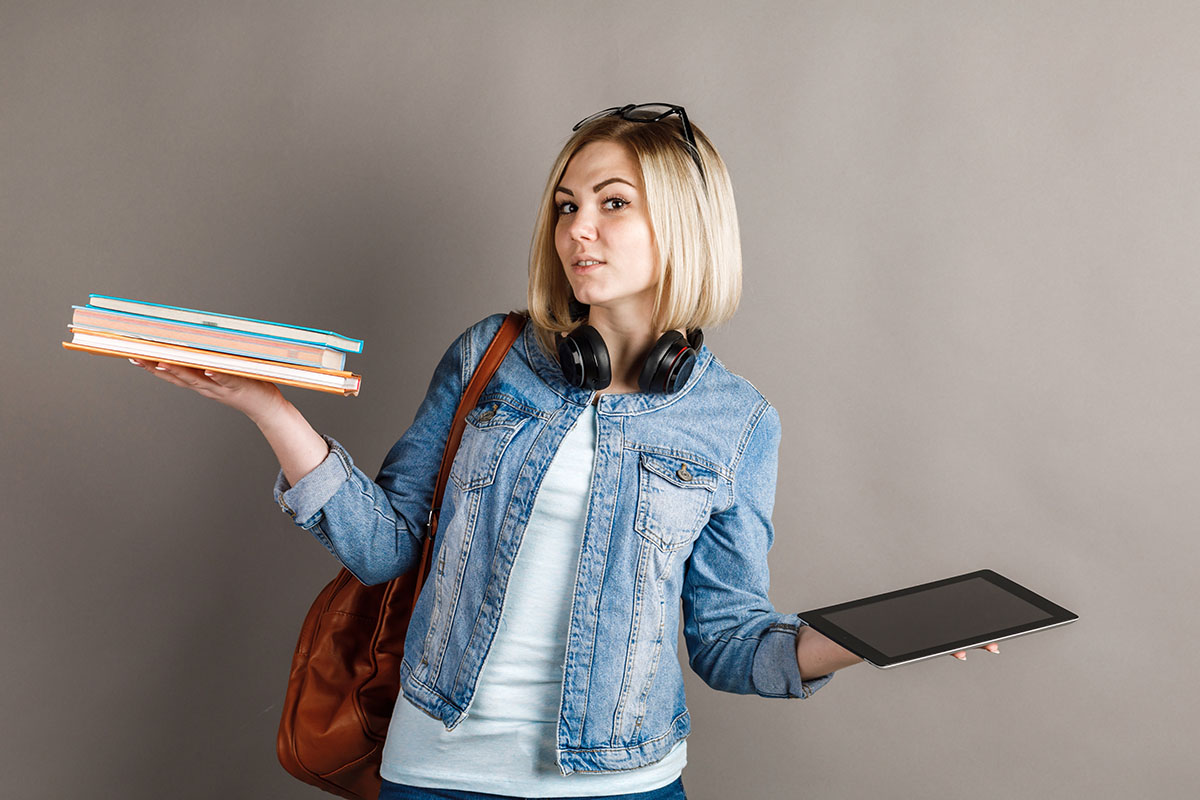 Book vs. e-book. Girl student holding a traditional textbook and reading e-books isolated on gray background. Choose between paper books and e-learning gadgets. The concept of modern education.