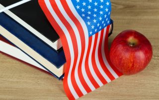American flag atop a stack of books, an apple to the right, view from above, American spelling versus british